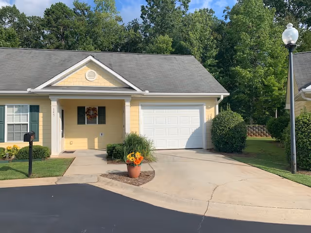 Exterior view of a single-story yellow house with white trim, a front porch decorated with a wreath, a white garage door, a driveway, and a flower pot with orange and yellow flowers. There is a black mailbox on the left and a street lamp on the right, with green trees in the background.