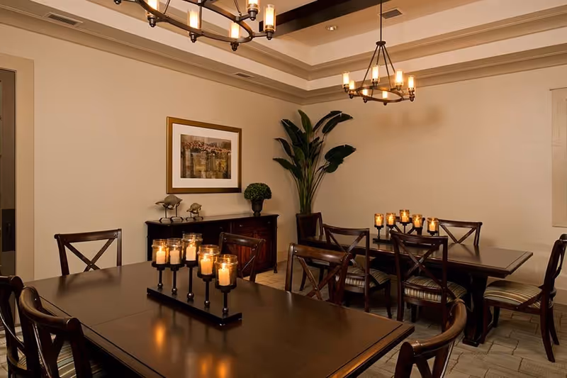 A warmly lit dining room with two dark wooden tables, each surrounded by wooden chairs with striped cushions. Each table has a centerpiece with multiple lit candles. The room features beige walls, a framed picture, a large green plant in the corner, and two chandeliers hanging from the ceiling.