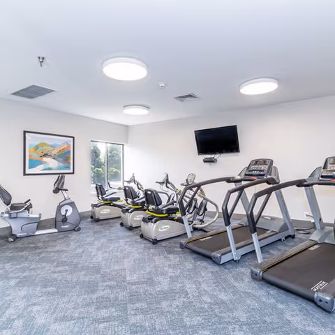 A fitness room with exercise equipment including treadmills, stationary bikes, and recumbent bikes. The room has gray carpet flooring, white walls, a wall-mounted TV, and a framed landscape painting. Ceiling lights provide bright illumination.