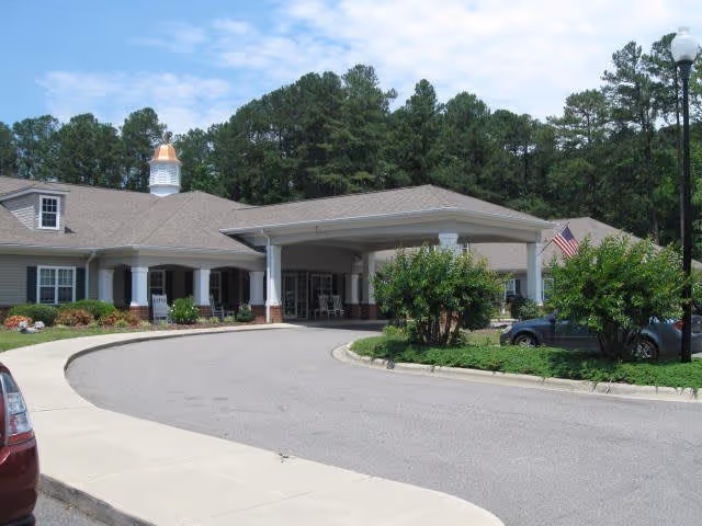 Front exterior view of Tara Plantation of Carthage facility showing a covered entrance with white columns, a circular driveway, landscaped bushes, an American flag, and trees in the background under a partly cloudy sky.