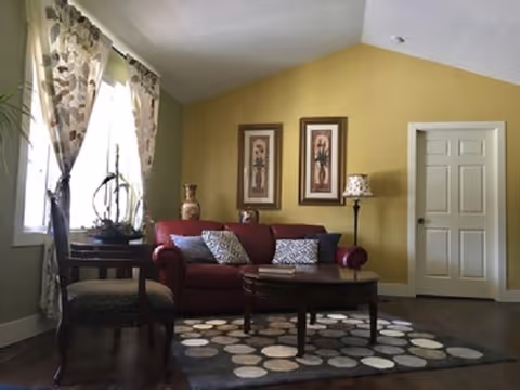 A cozy living room with a red sofa adorned with patterned cushions, a wooden coffee table on a circular patterned rug, two framed floral artworks on a yellow wall, a floor lamp with a patterned shade, and a wooden chair next to a window with floral curtains.
