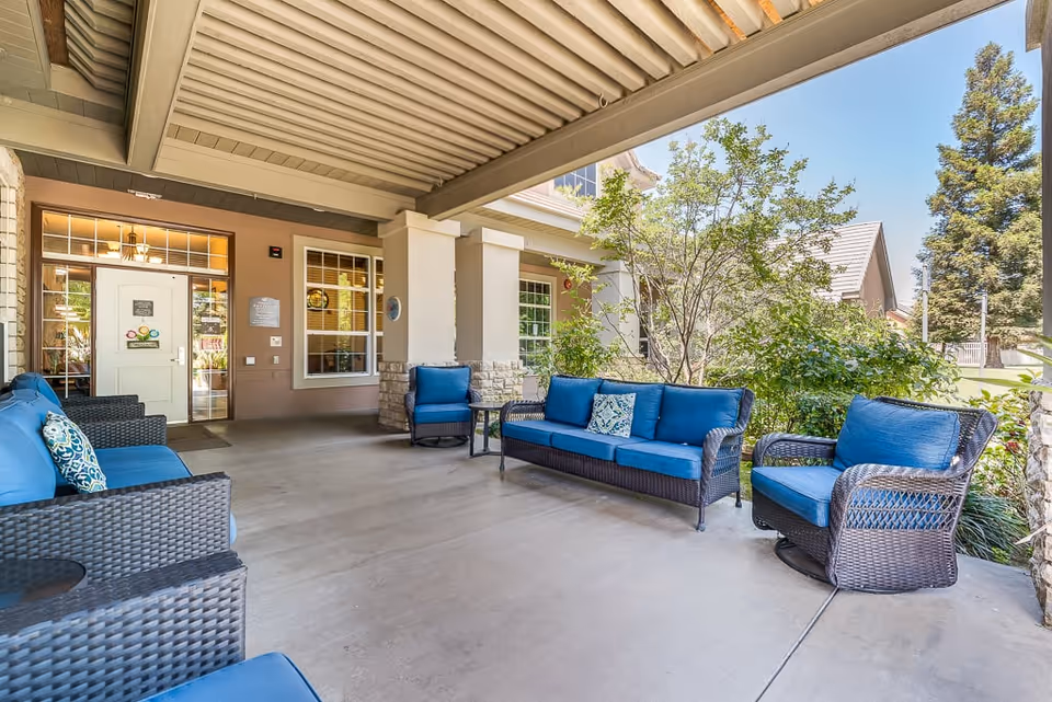 Covered outdoor patio area at Riverstone Terrace Senior Living with wicker furniture including blue cushioned sofas and chairs, small tables, and greenery surrounding the space under a beige roof structure.