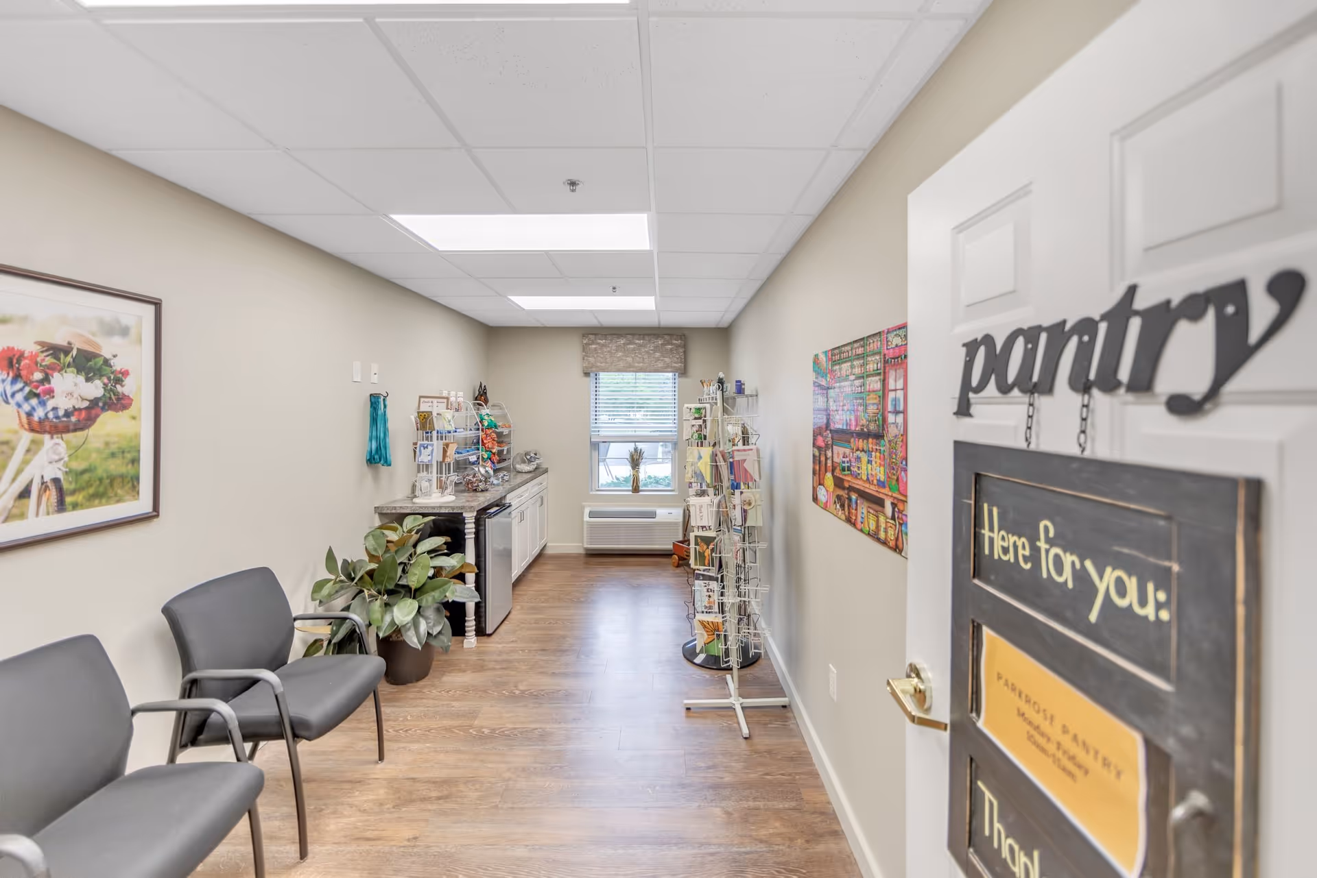Interior view of a pantry room at Parkrose Estates with a door labeled 'pantry' and a sign that says 'Here for you'. The room has wooden flooring, beige walls, a window with a blind, two gray chairs, a potted plant, a countertop with a mini fridge, and rotating racks holding various items.