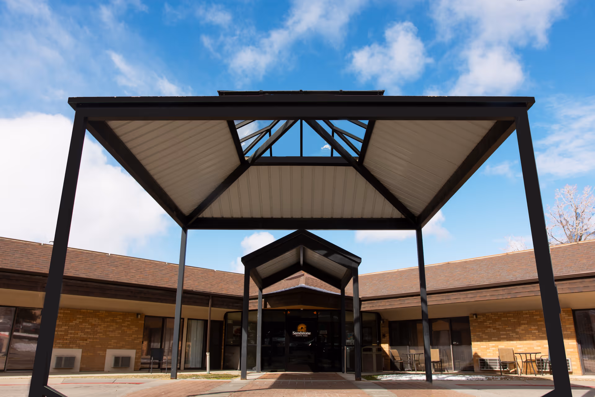 Entrance of Sandstone North Park facility with a covered walkway supported by black metal beams and a clear blue sky with some clouds in the background. The building has a brown roof and brick walls with glass doors at the entrance.