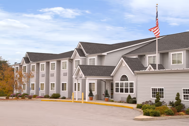 Exterior view of a two-story senior living facility building with gray siding, multiple windows, and a main entrance with a small covered porch. An American flag is flying on a flagpole near the entrance, and there are landscaped bushes and plants around the building.