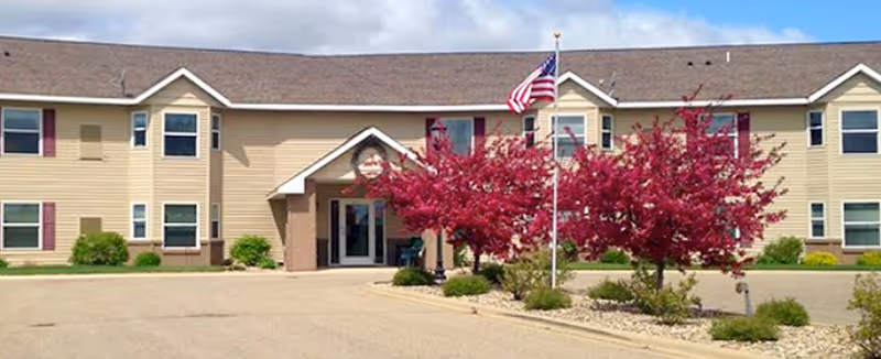 Front entrance of a two-story beige senior living building with a flagpole and blooming pink trees in the driveway.