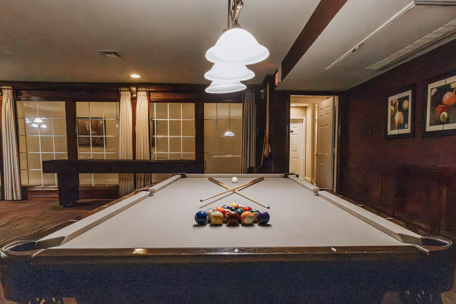 Interior view of a game room with a pool table in the center. The pool table has a white felt surface with billiard balls racked and two pool cues crossed on top. Above the table, three pendant lights hang from the ceiling. The room has large windows with curtains, dark wood paneling, and framed pictures on the wall.