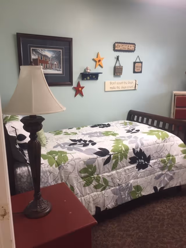 A small bedroom with a single bed covered in a white, green, black, and gray leaf-patterned quilt. A dark wooden headboard and footboard frame the bed. On the left side, there is a red nightstand with a decorative lamp. The wall behind the bed is light blue and decorated with framed artwork, wooden stars, and inspirational plaques.