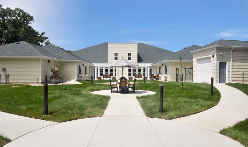 Outdoor courtyard area of Birchwood Cottages featuring a circular concrete pathway surrounding a grassy lawn with a table and chairs under a large umbrella. The courtyard is enclosed by single-story beige buildings with green roofs under a clear blue sky.
