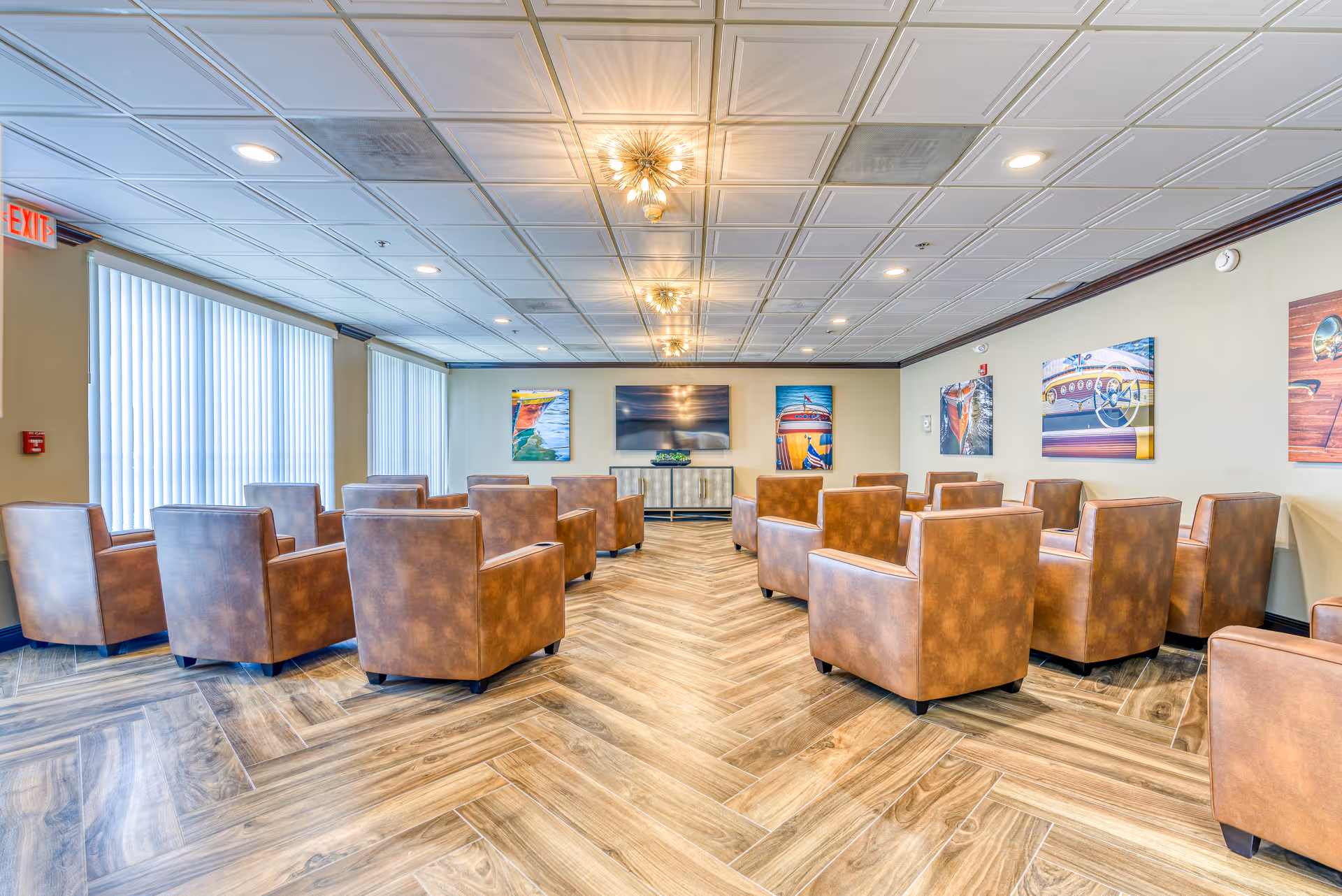 A bright communal lounge with brown leather armchairs arranged in rows facing a wall-mounted TV and colorful artwork.