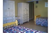 A bedroom with two wooden beds featuring floral patterned bedspreads. There is a white wooden dresser and a matching desk against a beige wall. The floor is tiled and the room has a simple, clean appearance.