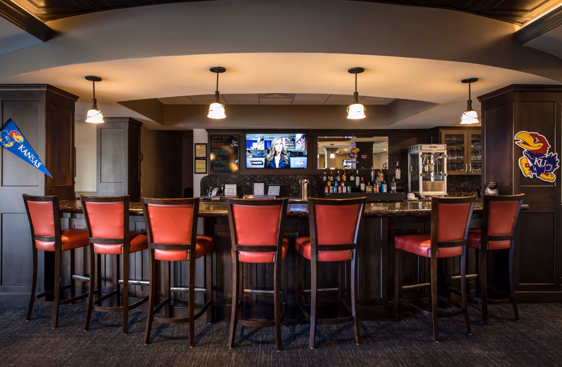 Interior bar/lounge with a row of red-cushioned bar stools at a dark wood counter, pendant lights, a TV, and Kansas Jayhawks decor.