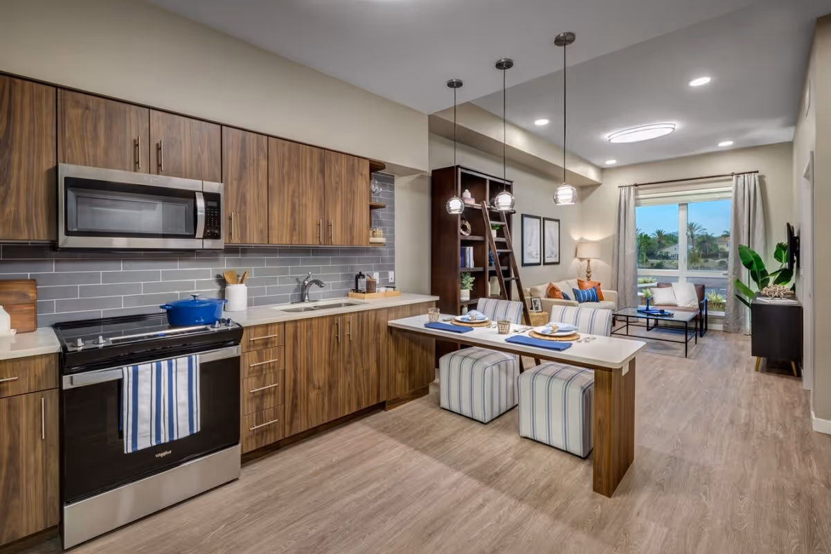Open-plan modern kitchen and living area with wood cabinets, a countertop dining peninsula with striped stools, and a seating area by a window.