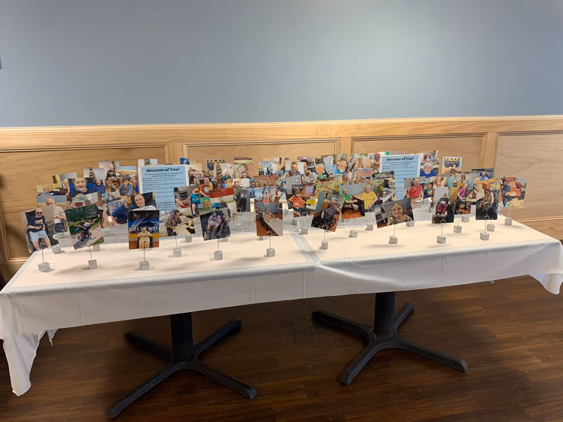 Two tables covered with white tablecloths displaying numerous small photo cards on stands, each featuring pictures of smiling elderly individuals and caregivers, set against a wooden paneled wall with informational posters titled 'Because of You!'