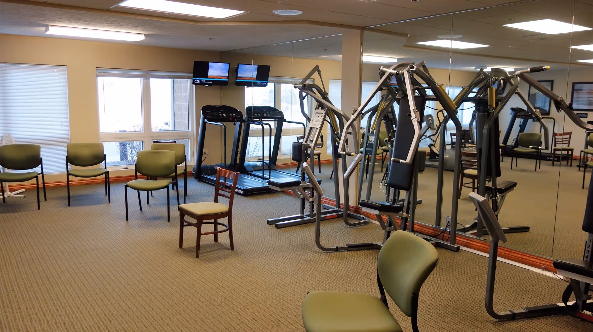 A fitness room in a senior living facility with exercise equipment including two treadmills and a multi-station weight machine. Several green chairs and one wooden chair are arranged around the room. Large windows and a wall of mirrors reflect the equipment and chairs.
