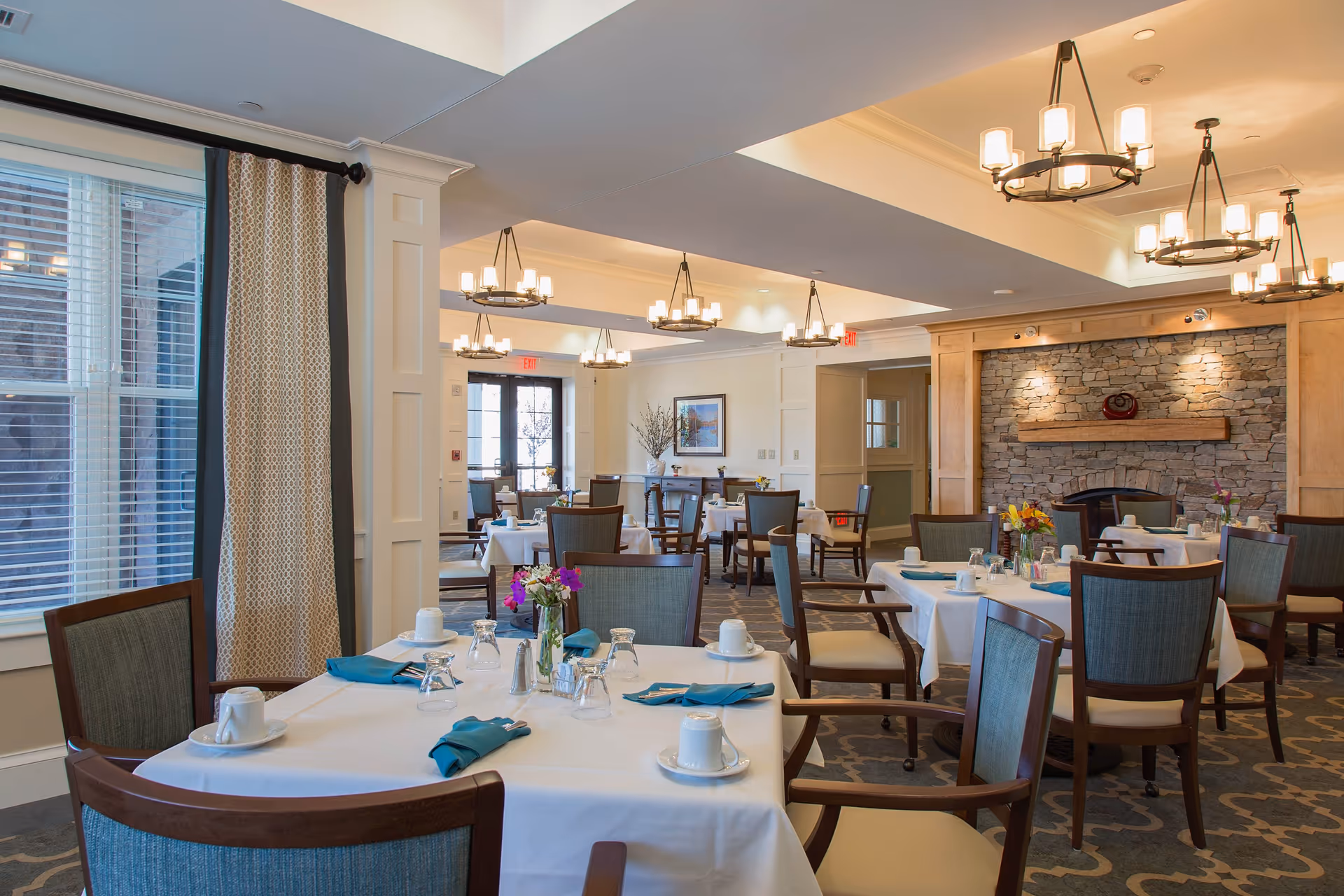 A well-lit dining room with multiple tables covered in white tablecloths, each set with cups, glasses, napkins, and small flower arrangements. The room features wooden chairs with cushioned seats, a stone fireplace with a wooden mantle, and several modern chandeliers hanging from the ceiling. Large windows with curtains allow natural light to enter the space.
