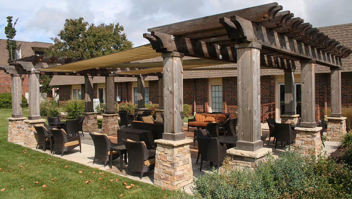 Outdoor seating area at Foxwood Springs Senior Living featuring a wooden pergola with stone pillars, multiple wicker chairs and tables, and a cushioned bench. The area is surrounded by grass and shrubbery with a brick building in the background.