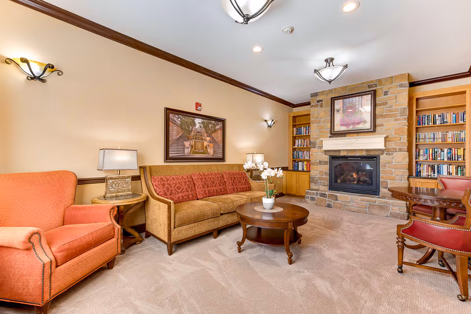 A cozy senior living room featuring a stone fireplace with a framed picture above it, built-in wooden bookshelves filled with books on either side, a beige sofa with red patterned cushions, an orange armchair, a round wooden coffee table with a white orchid plant, and a wooden side table with a lamp. The room has beige walls with dark wood trim and soft carpeted flooring.