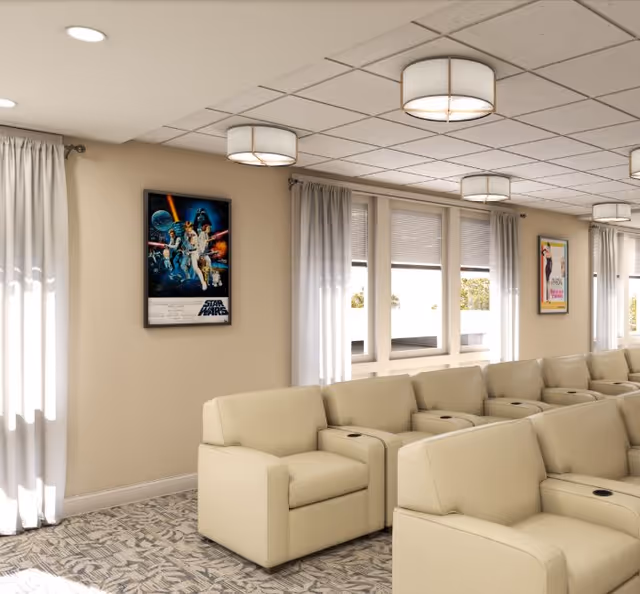 A bright, modern lounge area with beige armchairs arranged in rows, each featuring built-in cup holders. The room has large windows with white curtains allowing natural light to enter. The walls are decorated with framed movie posters, including one for Star Wars. The ceiling has multiple round light fixtures.