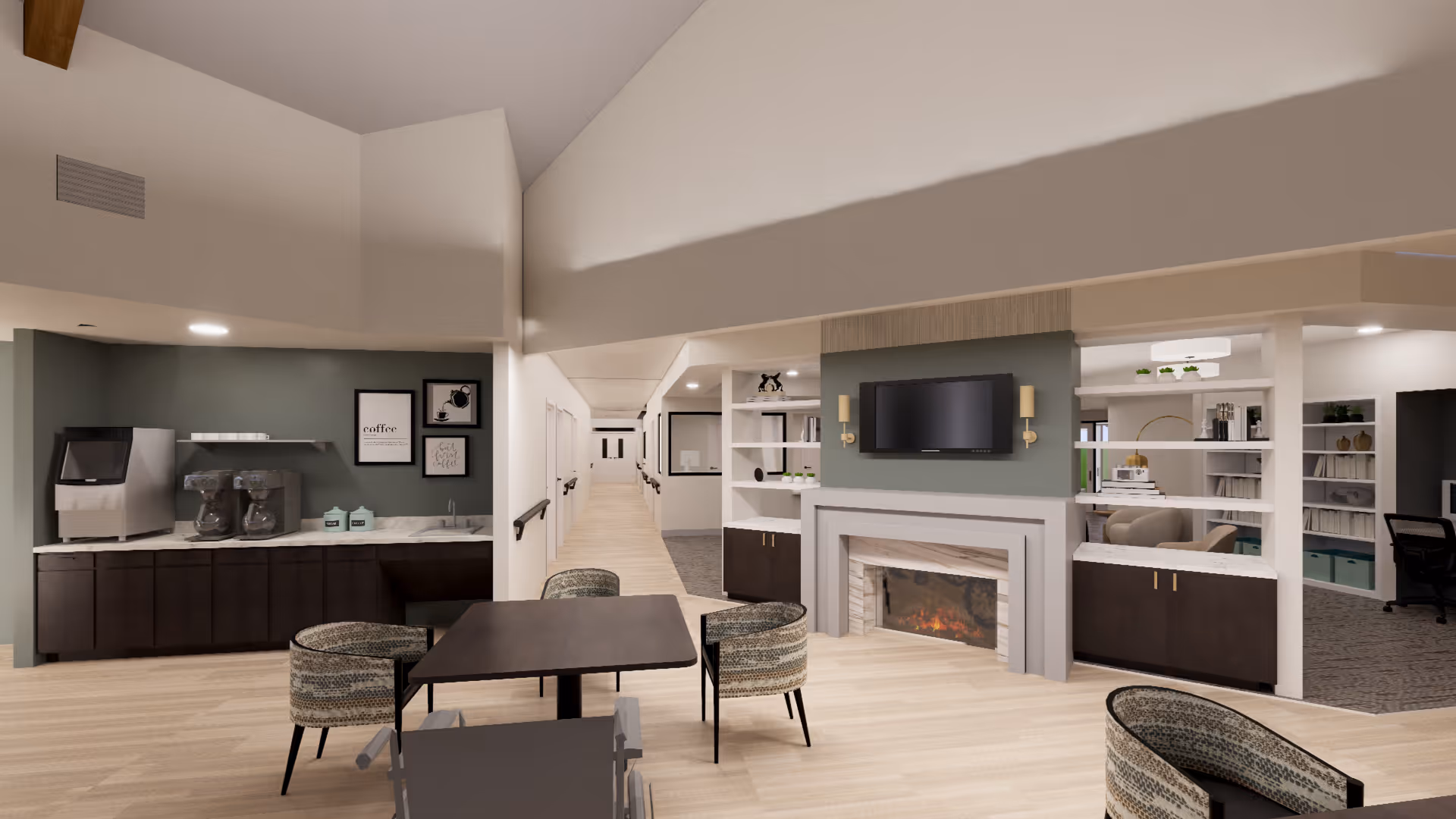 A modern senior living facility common area featuring a dark wood table with four patterned chairs, a fireplace with a mounted TV above it, and a coffee station with two coffee makers and a water dispenser against a green accent wall. The space has light wood flooring, neutral walls, and an open layout with a hallway leading to other rooms.