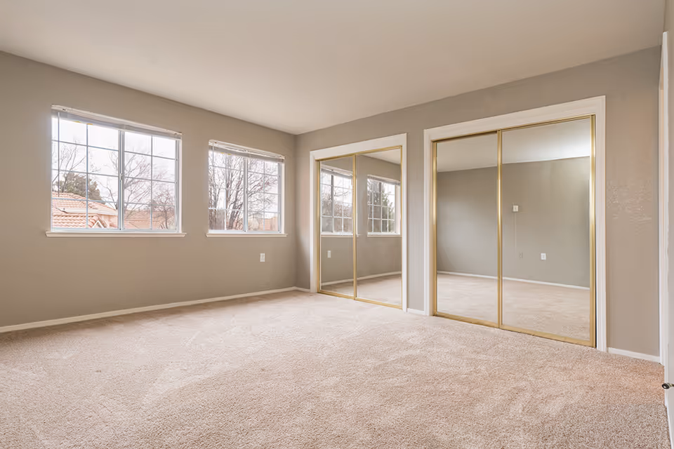Empty bedroom with beige carpet, two large windows with white frames, and two mirrored sliding closet doors with gold trim. The walls are painted light gray.