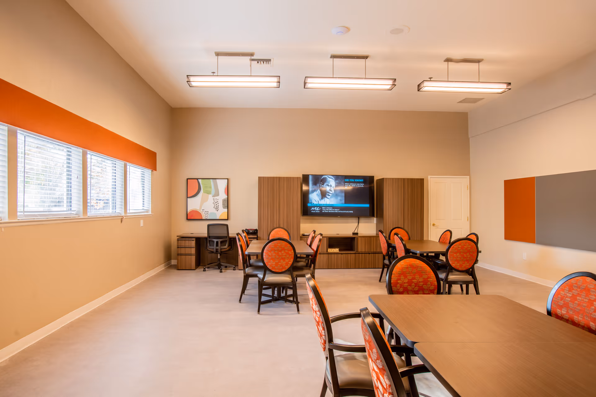 A spacious, well-lit common room with multiple tables and chairs arranged for group seating. The chairs have orange patterned upholstery. On the far wall, there is a flat-screen TV mounted between two wooden cabinets. A desk with a chair is positioned near the window on the left side of the room. The walls are painted beige with orange and gray accent panels. Large windows on the left wall allow natural light to enter the room.