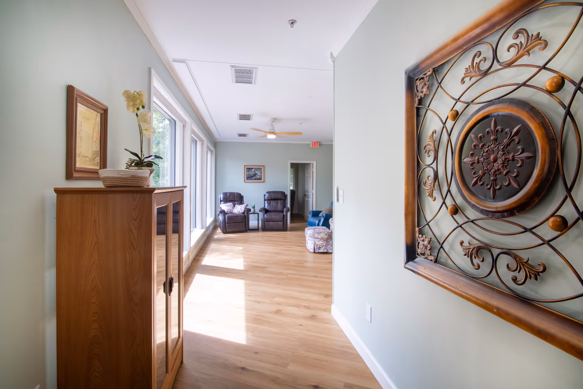 A bright hallway with light wooden flooring and pale green walls. On the left side, there is a wooden cabinet with a potted orchid and a framed picture above it. At the end of the hallway, there are two dark reclining chairs with a small table between them, and a framed picture on the far wall. On the right wall, there is a decorative metal wall art piece with intricate patterns. Large windows on the left side let in natural light.