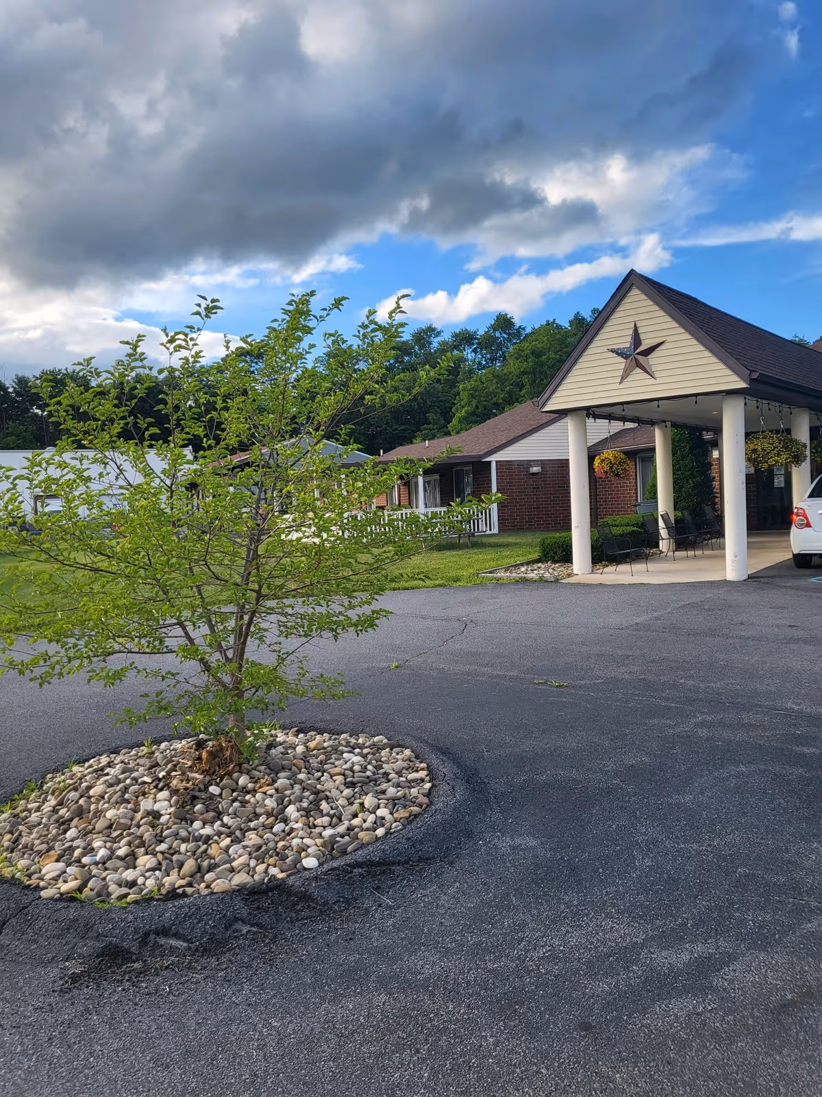 Outdoor view of a senior living facility entrance with a small tree planted in a circular bed of rocks in the foreground. The building has a covered entrance supported by white columns, with hanging flower baskets and a large decorative star on the gable. A white car is partially visible parked near the entrance. The sky is partly cloudy with patches of blue.