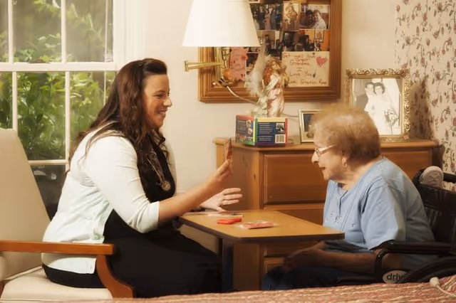 A young female caregiver or nurse is sitting and smiling while interacting with an elderly woman in a wheelchair across a small table in a warmly decorated room with family photos and a lamp in the background.