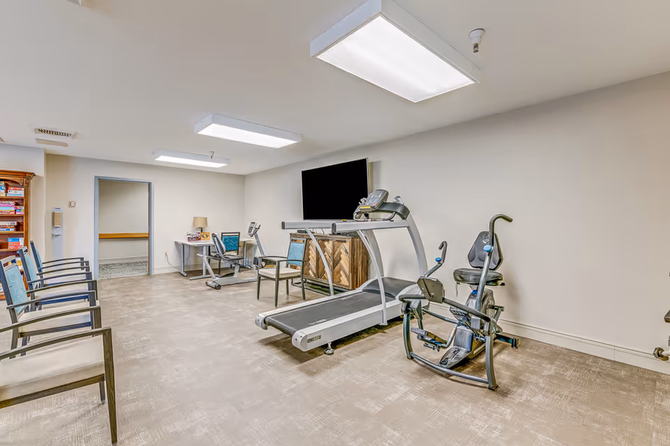 Indoor exercise room with a treadmill, recumbent bike, chairs and a wall-mounted TV in a senior living facility.