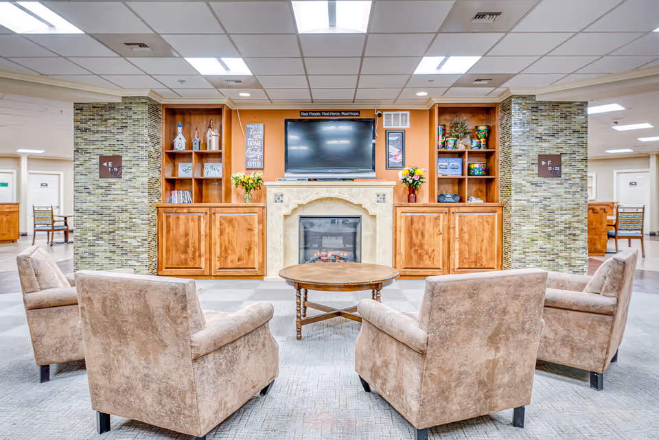 Community lounge with four upholstered chairs around a round coffee table facing a fireplace and wall-mounted TV flanked by wooden cabinets and shelves.