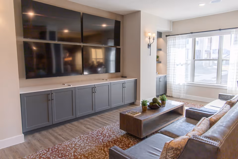 A modern living room with a large wall-mounted entertainment system consisting of four flat-screen TVs above a long gray cabinet. The room features a wooden coffee table with decorative plants, a patterned area rug, a leather sofa with cushions, and a large window with sheer white curtains letting in natural light.
