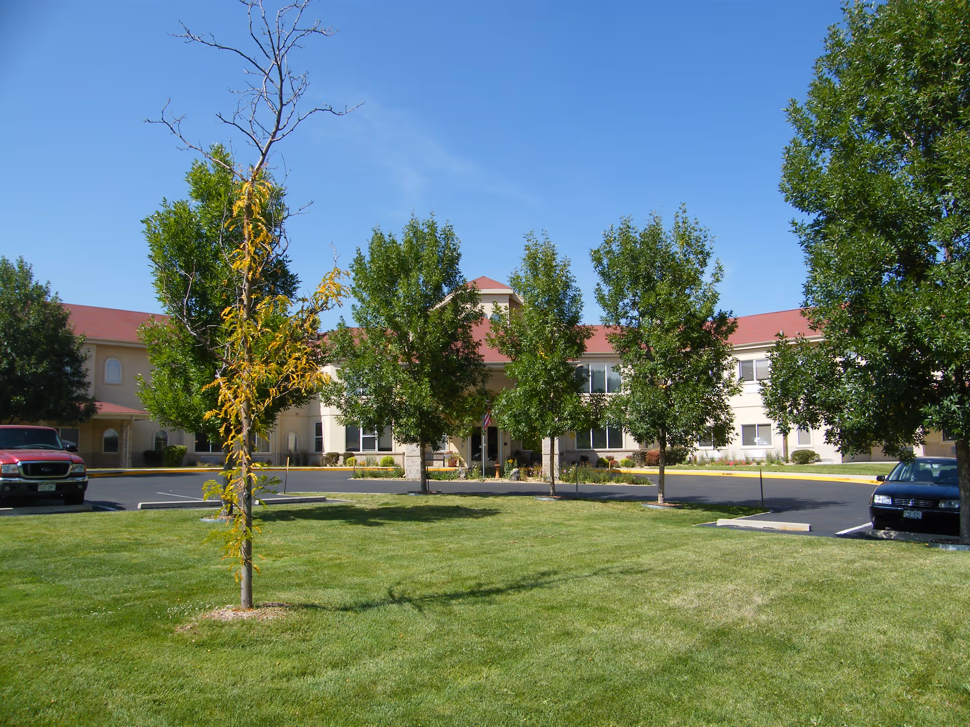 Exterior view of Marycrest Assisted Living facility showing a two-story building with a red roof, surrounded by green trees and a well-maintained lawn under a clear blue sky. There are parked cars visible on either side of the building.
