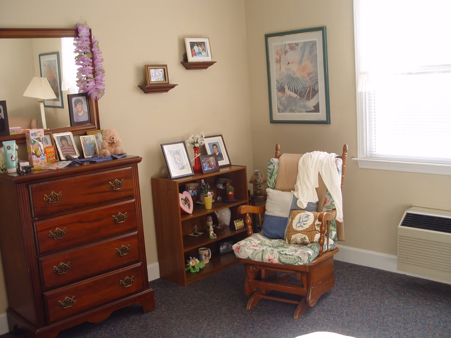 A cozy corner of a room featuring a wooden rocking chair with floral cushions and several pillows, a wooden dresser with a mirror adorned with a purple lei, and a small wooden bookshelf displaying framed photos and decorative items. A framed flamingo artwork hangs on the wall next to a window with white blinds, and a wall-mounted air conditioning unit is below the window.