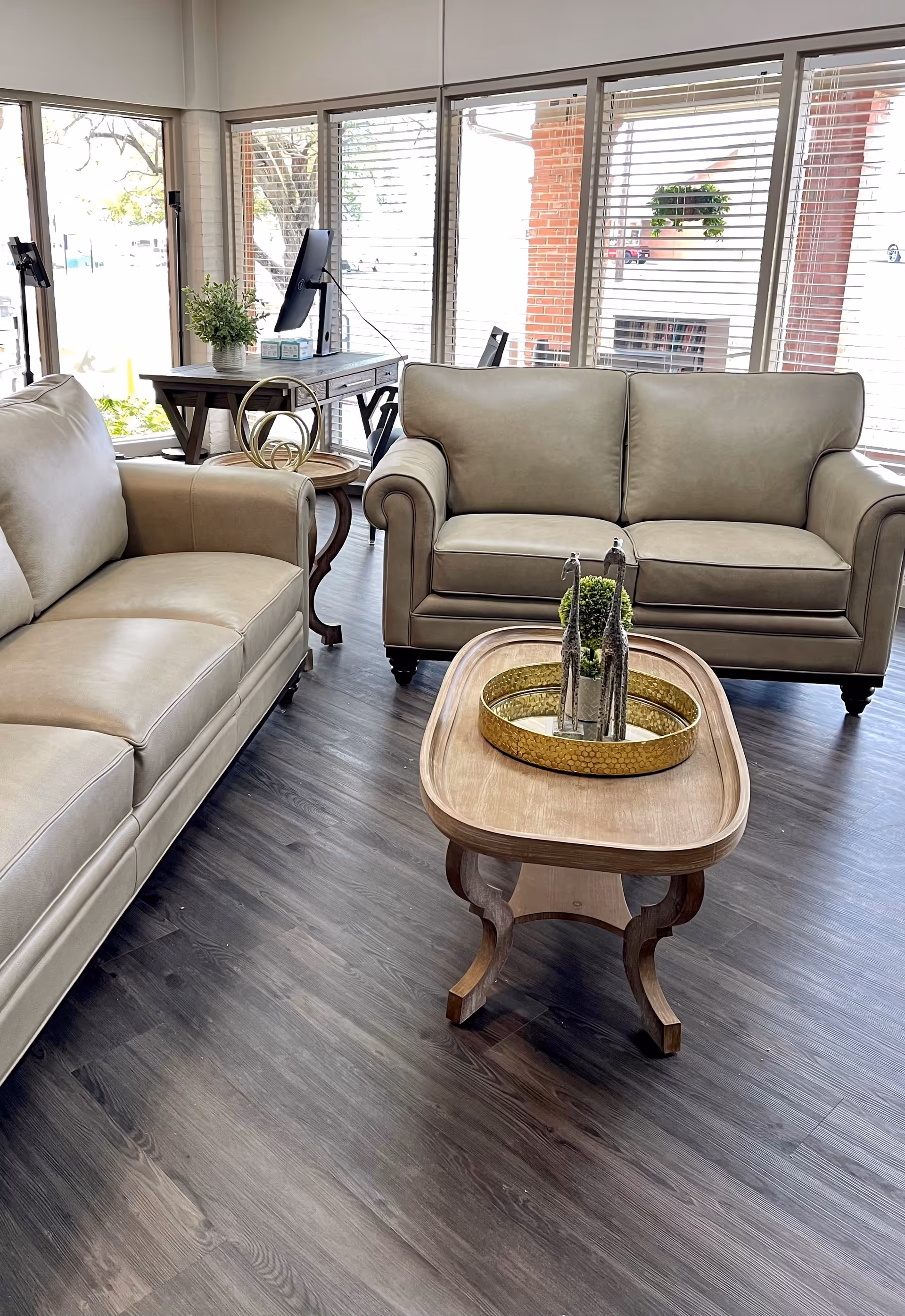 A bright and modern living room area with two beige leather sofas facing each other, a wooden coffee table with decorative items on it, and a side table with a plant and a computer monitor near large windows with blinds.