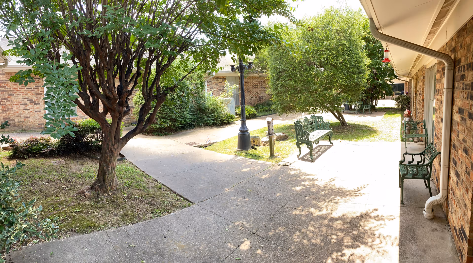 Sunny enclosed courtyard with paved walkways, trees, green metal benches, a lamp post, and a brick building exterior.
