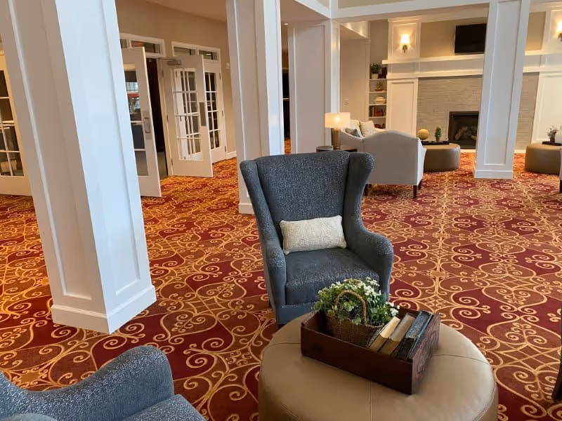 A spacious living room area with patterned red and gold carpet, white pillars, and multiple seating arrangements including gray armchairs and beige ottomans. A small wooden tray with books and a potted plant is placed on one ottoman. The room features a fireplace with a TV mounted above it and built-in shelves on either side.