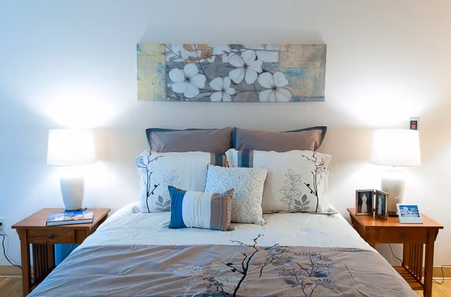 A neatly made bed with multiple pillows and a floral patterned comforter. Two wooden nightstands flank the bed, each with a lamp. One nightstand has a few framed photos and a small card, while the other has a book. Above the bed hangs a rectangular piece of artwork featuring white flowers on a muted background.