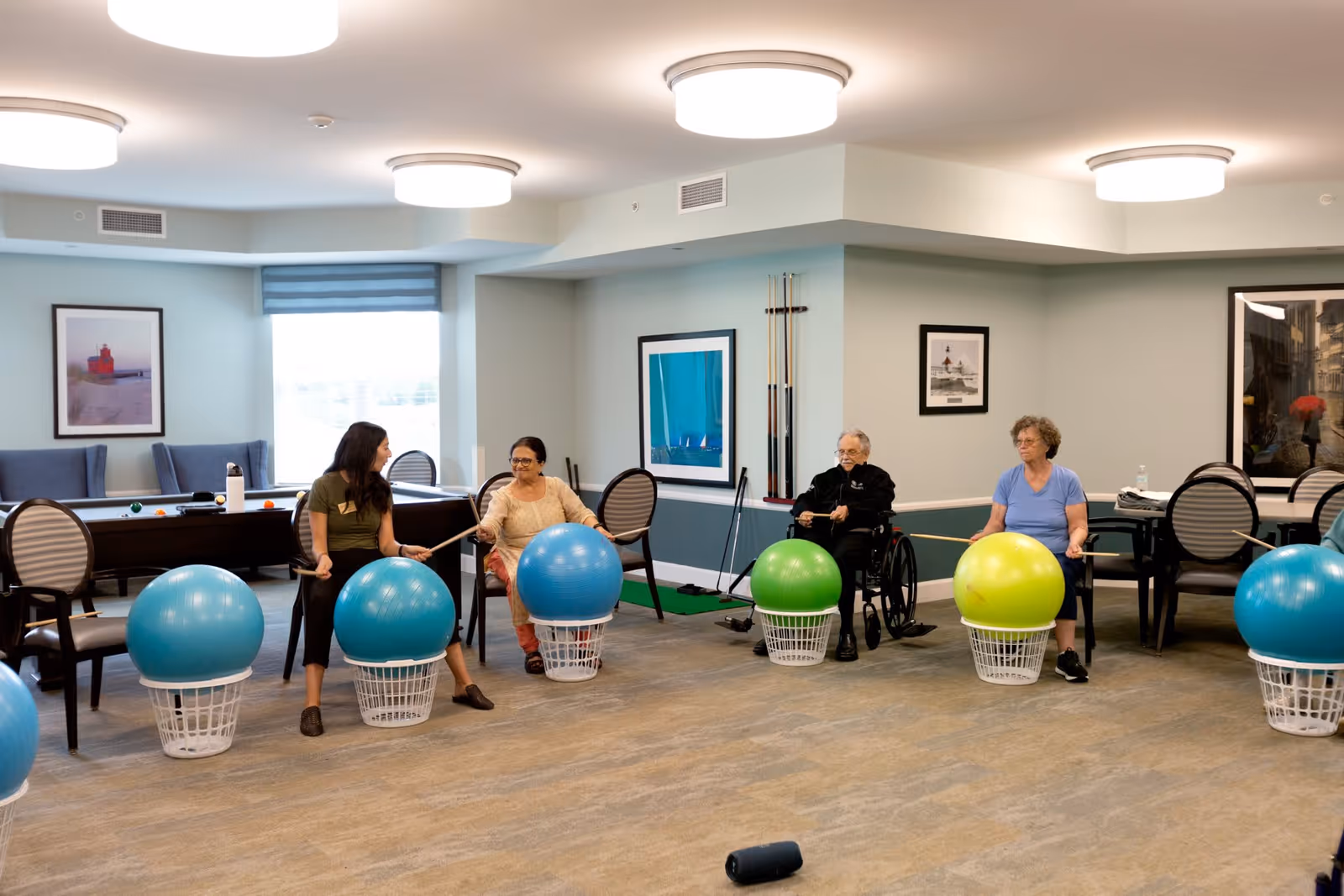 A group of four people sitting in a spacious room, each with a large exercise ball placed on a laundry basket in front of them. They are holding drumsticks and appear to be participating in a seated exercise or drumming activity. The room has light-colored walls, framed pictures, and several chairs and tables in the background.