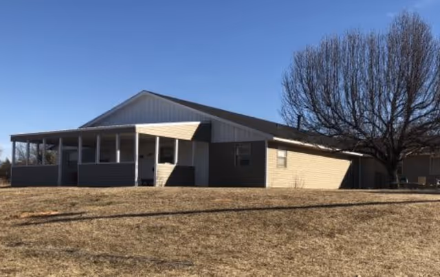 Single-story building with a covered porch and a large leafless tree beside it, situated on a dry grassy area under a clear blue sky.