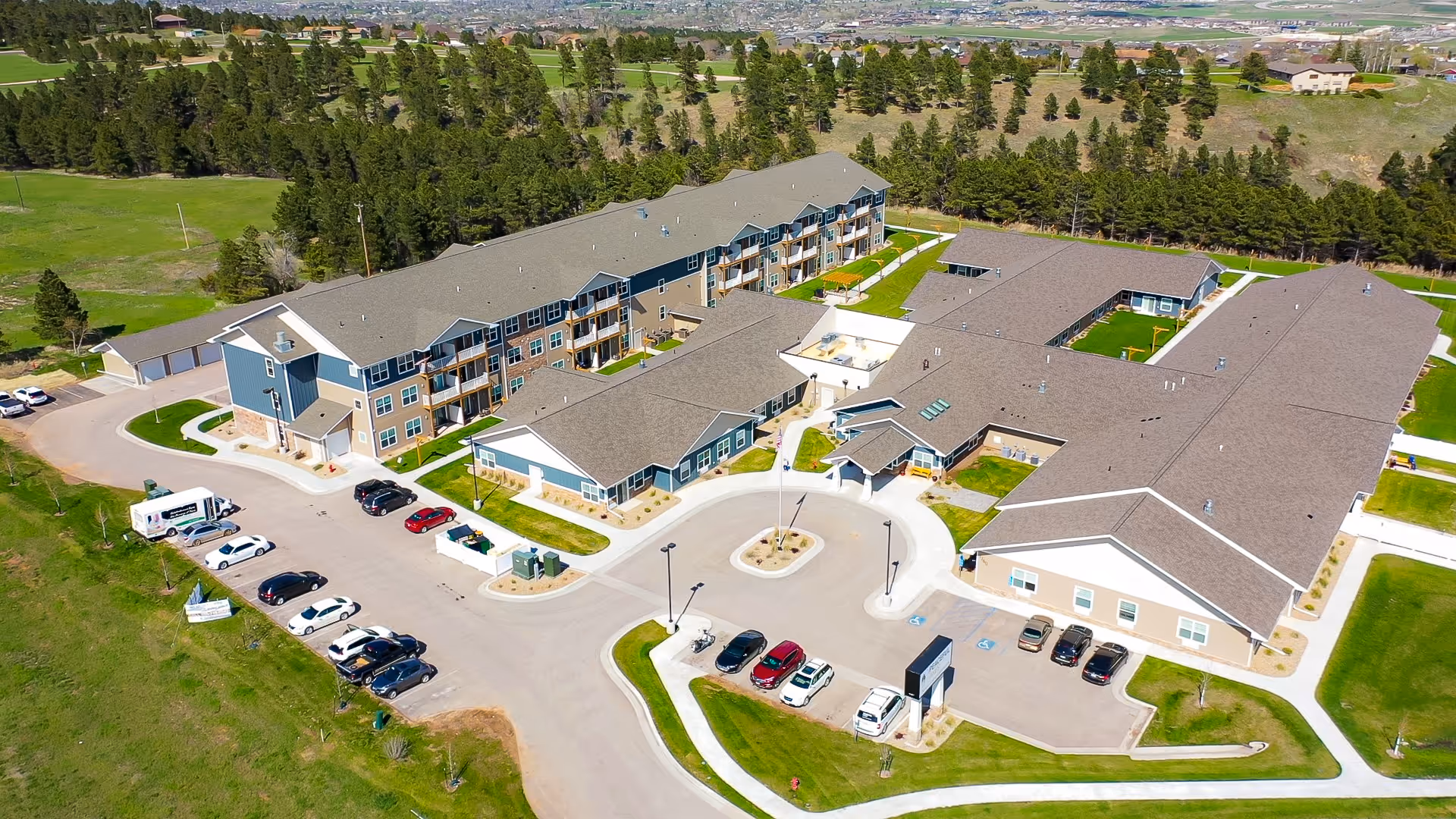 Aerial view of Peaceful Pines Senior Living at Rapid City showing multiple connected buildings with gray roofs and blue siding, surrounded by green lawns and trees. Several cars are parked in the parking lot, and the facility is situated in a semi-rural area with open fields and distant hills in the background.