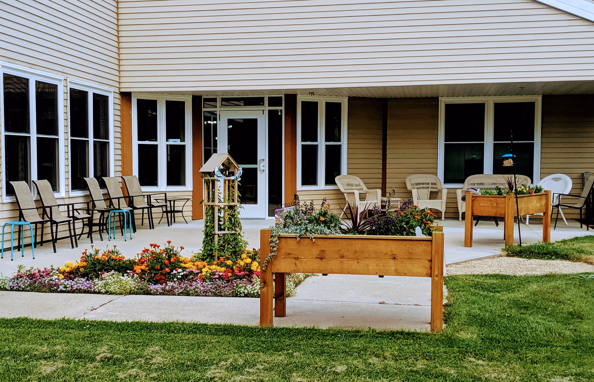 Outdoor patio area of a senior living facility with multiple chairs arranged along the building's wall and around small tables. There are raised wooden garden beds filled with plants and flowers, a flower bed with colorful flowers, and a small decorative structure in the center. The building has beige siding and multiple windows.