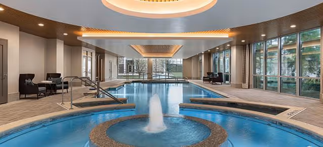 Indoor swimming pool area with a circular hot tub featuring a central water fountain in the foreground. The pool area has large floor-to-ceiling windows on the right side, letting in natural light and showing trees outside. The ceiling has modern recessed lighting and wooden panel accents. There are seating areas with chairs along the left side near the windows.