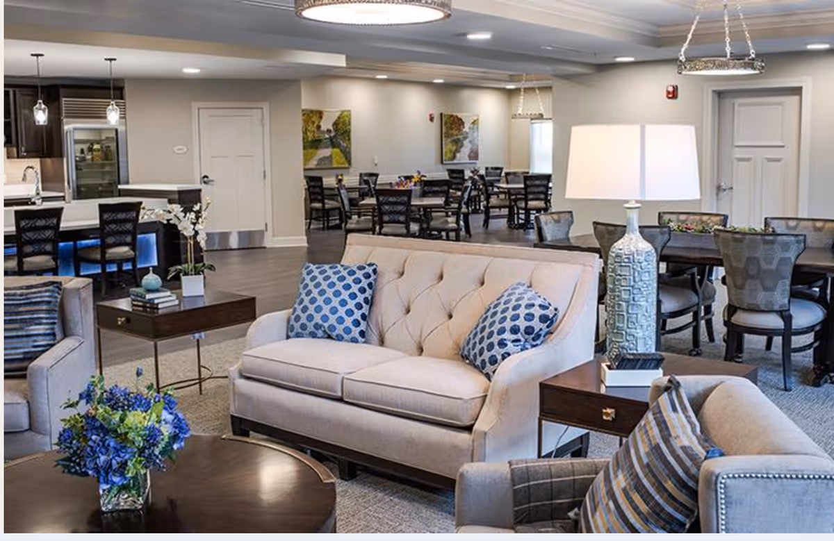 A spacious and well-lit common area in a senior living facility featuring beige sofas with blue patterned pillows, a round wooden coffee table with a floral arrangement, side tables with lamps, and multiple dining tables and chairs in the background. The room has a modern and cozy design with neutral tones and decorative wall art.