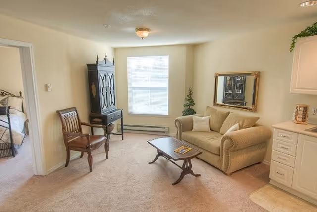 A cozy living room area with a beige couch, a wooden coffee table, a wooden chair, and a tall black cabinet near a window with blinds. There is a mirror on the wall above the couch and a small green plant in the corner. To the right, part of a kitchen counter and cabinets are visible. An adjoining bedroom with a bed is partially visible through an open doorway on the left.