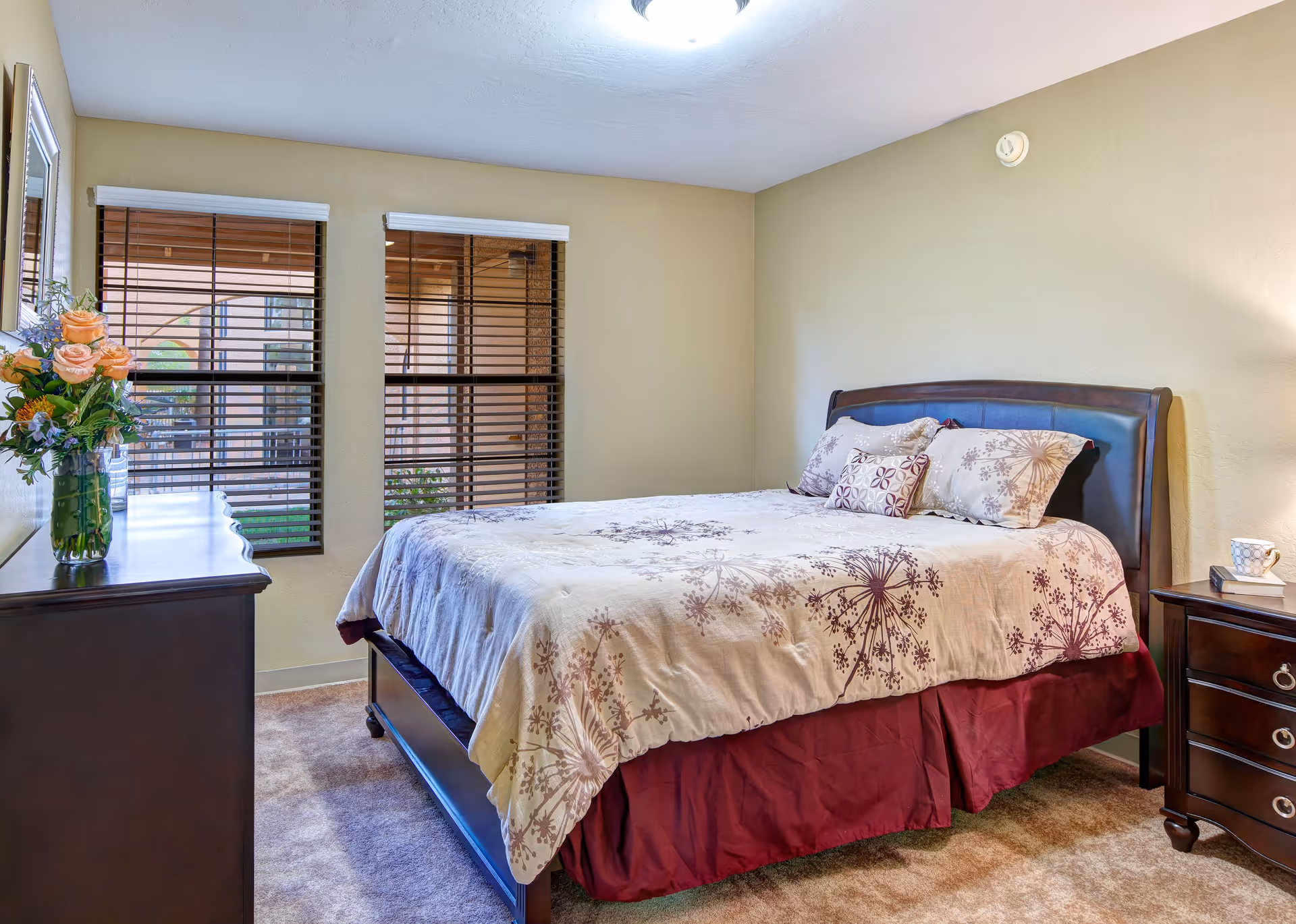 A cozy bedroom with a large bed featuring a floral patterned comforter and multiple pillows. The room has beige walls, two windows with wooden blinds, a dark wooden dresser with a vase of flowers, and a matching nightstand with a cup and book.
