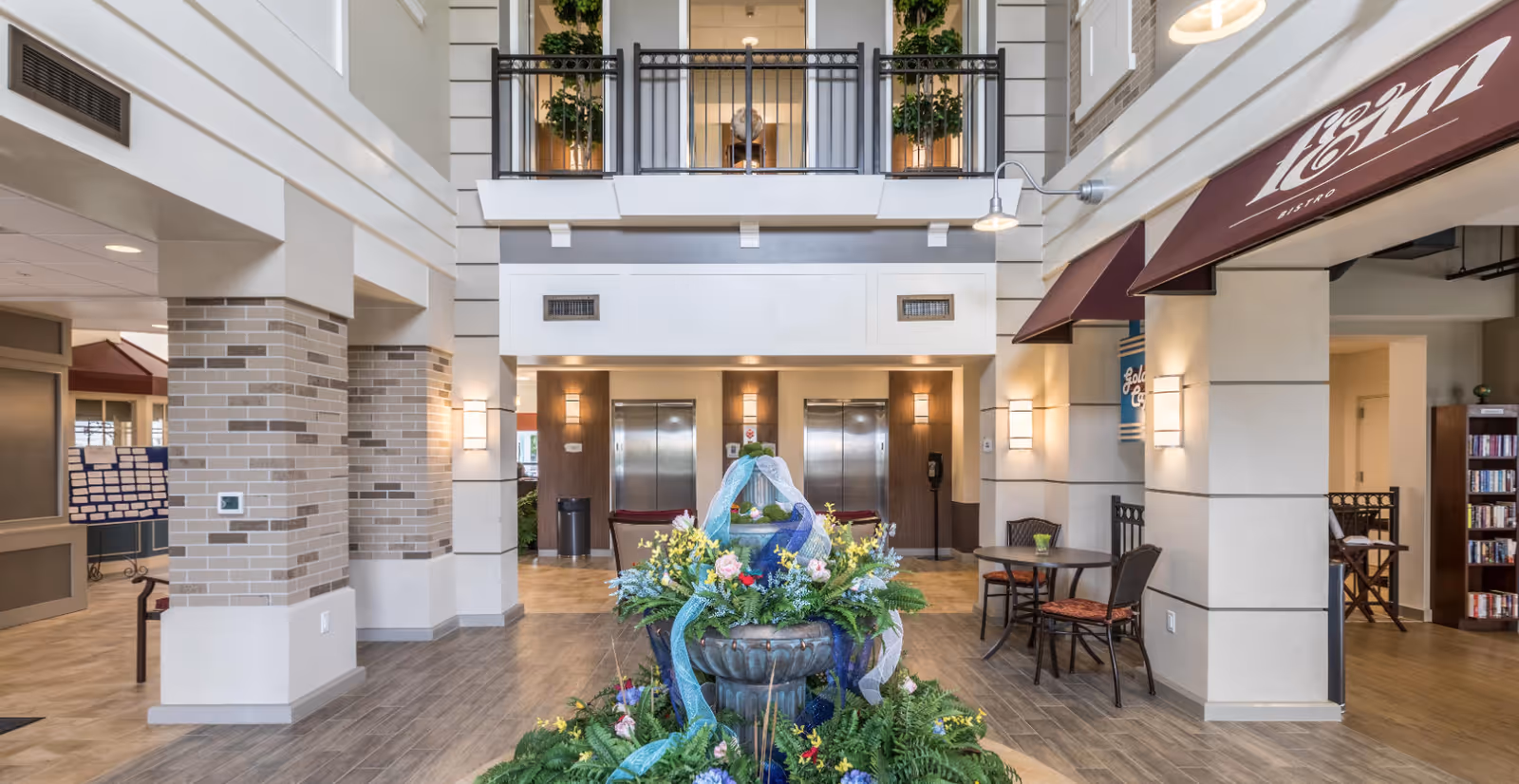 Interior view of a senior living facility lobby with a central decorative fountain adorned with flowers and blue ribbons. The space features two elevators in the background, a seating area with tables and chairs to the right, and a second-floor balcony with plants. The area has a modern design with light-colored walls, brick pillars, and wood flooring.