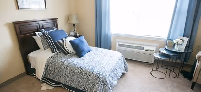 A neatly made bed with a wooden headboard, adorned with multiple pillows in shades of blue and white. Next to the bed is a small nightstand with a lamp and a clock. A large window with blue curtains lets in natural light, and below it is an air conditioning unit. To the right, there is a set of nested round tables with a framed photo and a small flower vase on top. The room has beige walls and carpeted flooring.