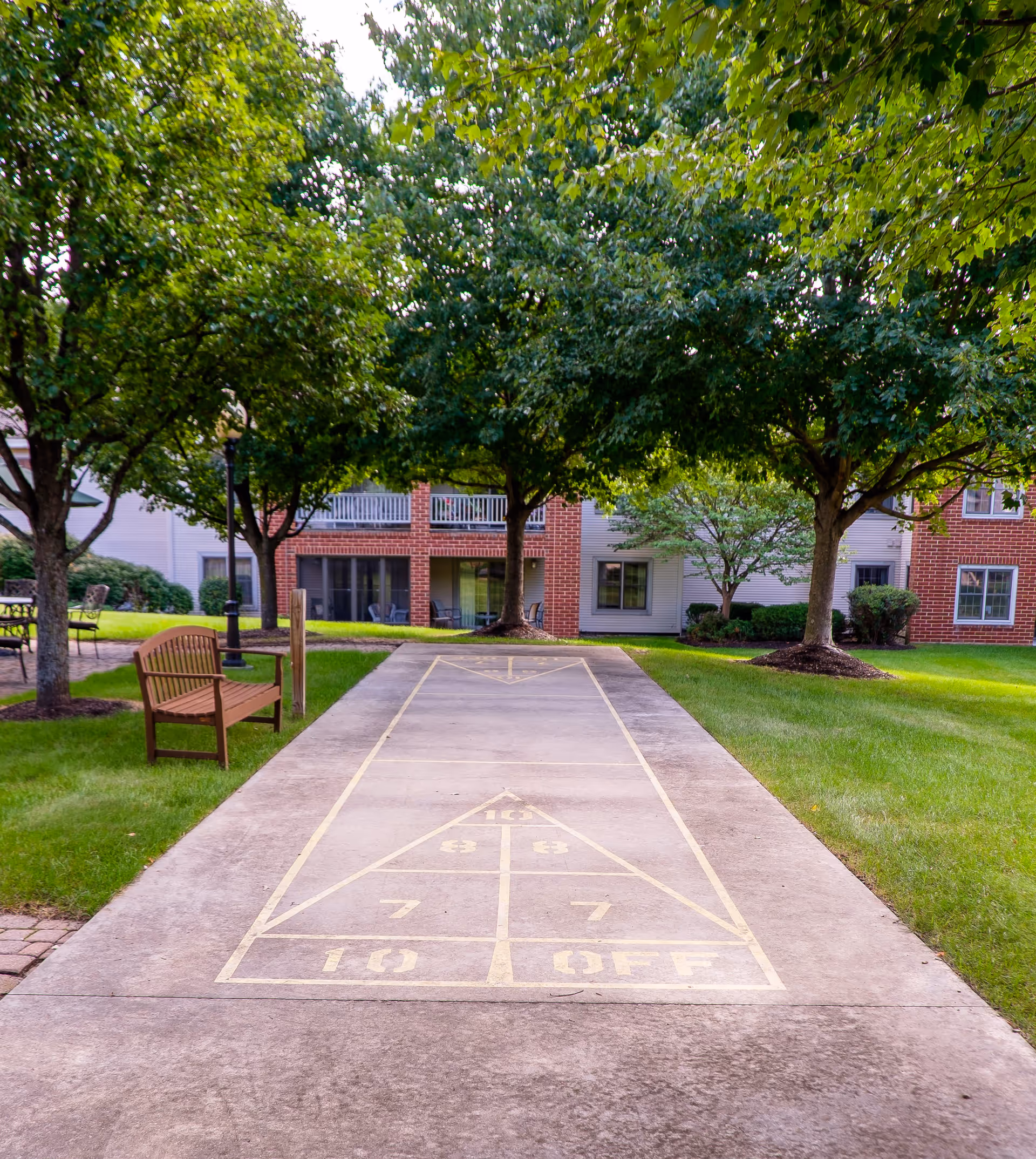 Outdoor shuffleboard court on a concrete pathway surrounded by green grass and trees, with a wooden bench on the left side and a brick building with balconies in the background.