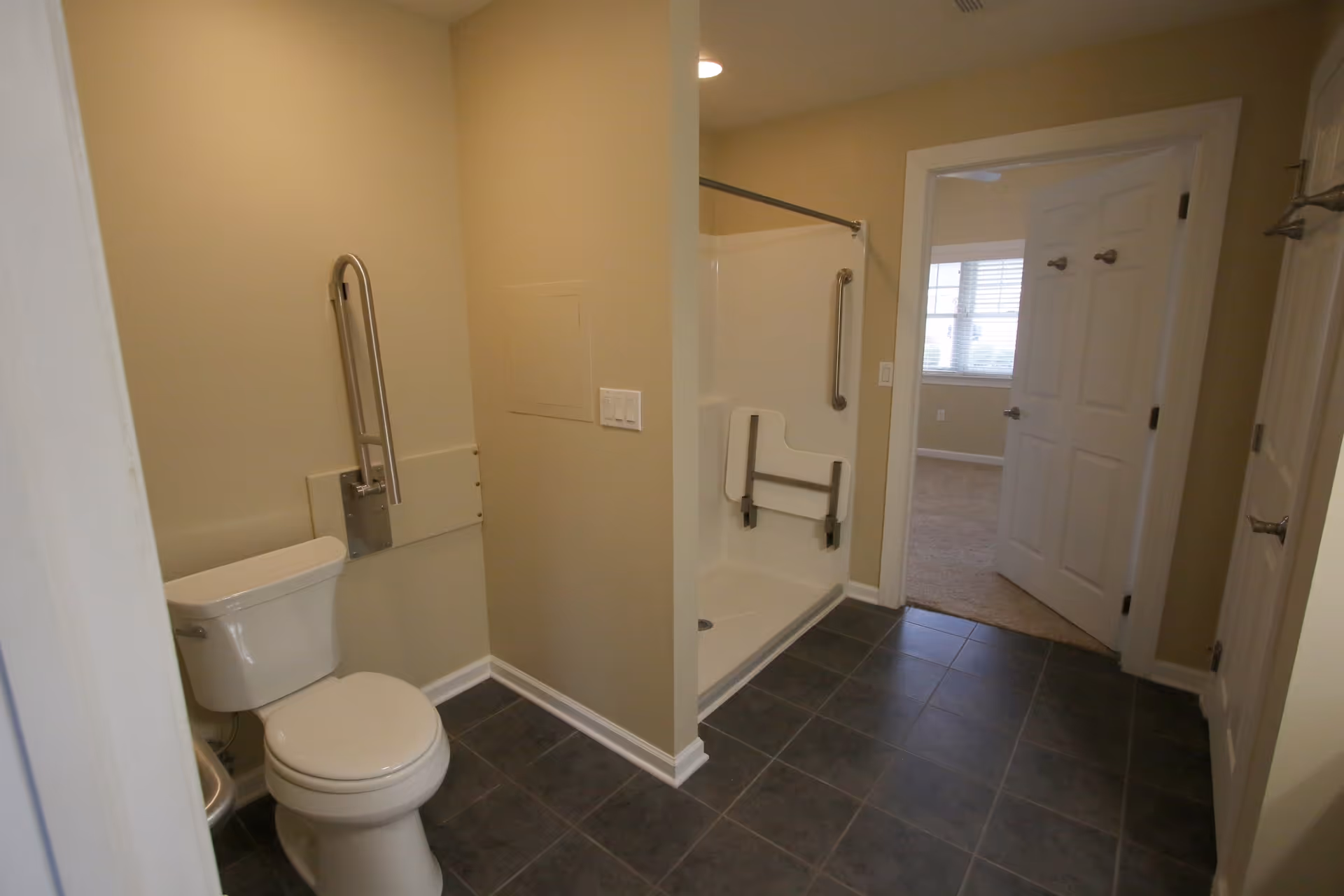 A bathroom with a white toilet, beige walls, and a walk-in shower with a fold-down seat and grab bars. The floor is tiled in dark gray, and there is an open door leading to a carpeted room with a window.